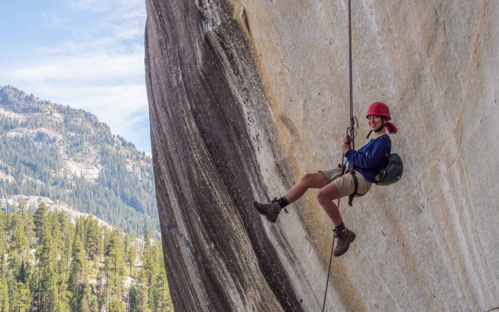 A person wearing safety gear is suspended by ropes mid air against the backdrop of a large smooth rock wall. There are evergreen trees, a mountain and blue skies in the background. 